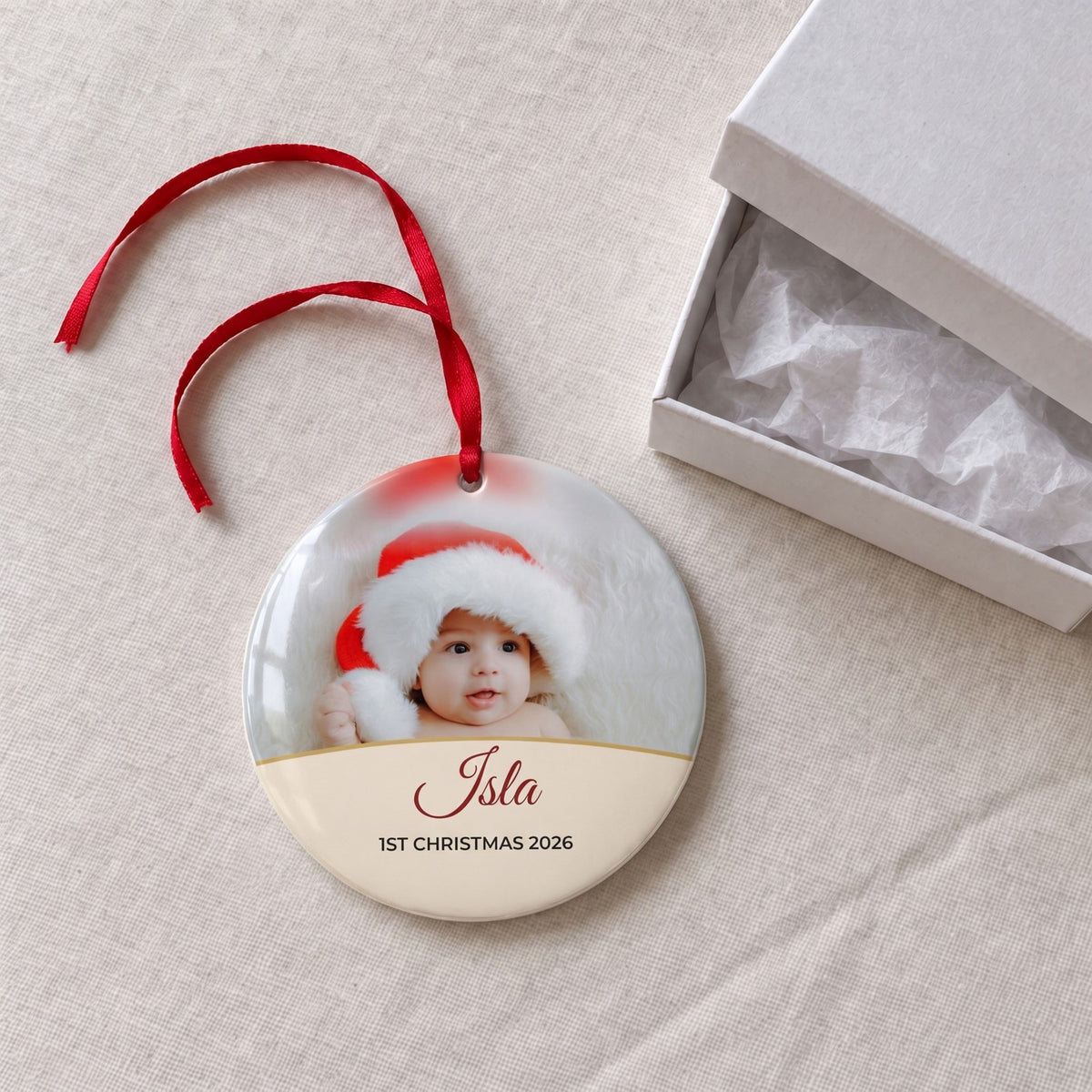 Cream personalised Baby's 1st Christmas photo bauble on a tablecloth beside a white gift box