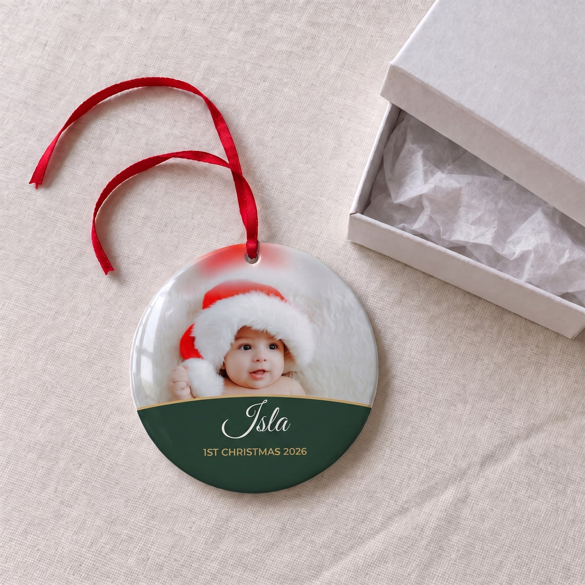 Forest green personalised Baby's 1st Christmas photo bauble on a tablecloth beside a white gift box