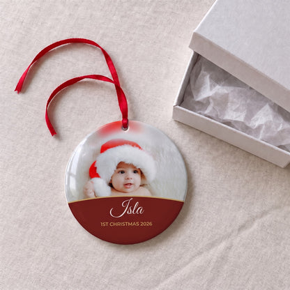 Berry red personalised Baby's 1st Christmas photo bauble on a tablecloth beside a white gift box